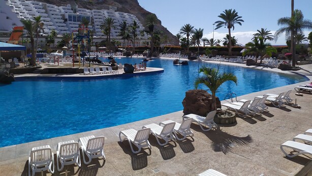 A resort pool with lounge chairs and palm trees. - Amazing ocean view apartment in Taurito, Mogán (Mogan)