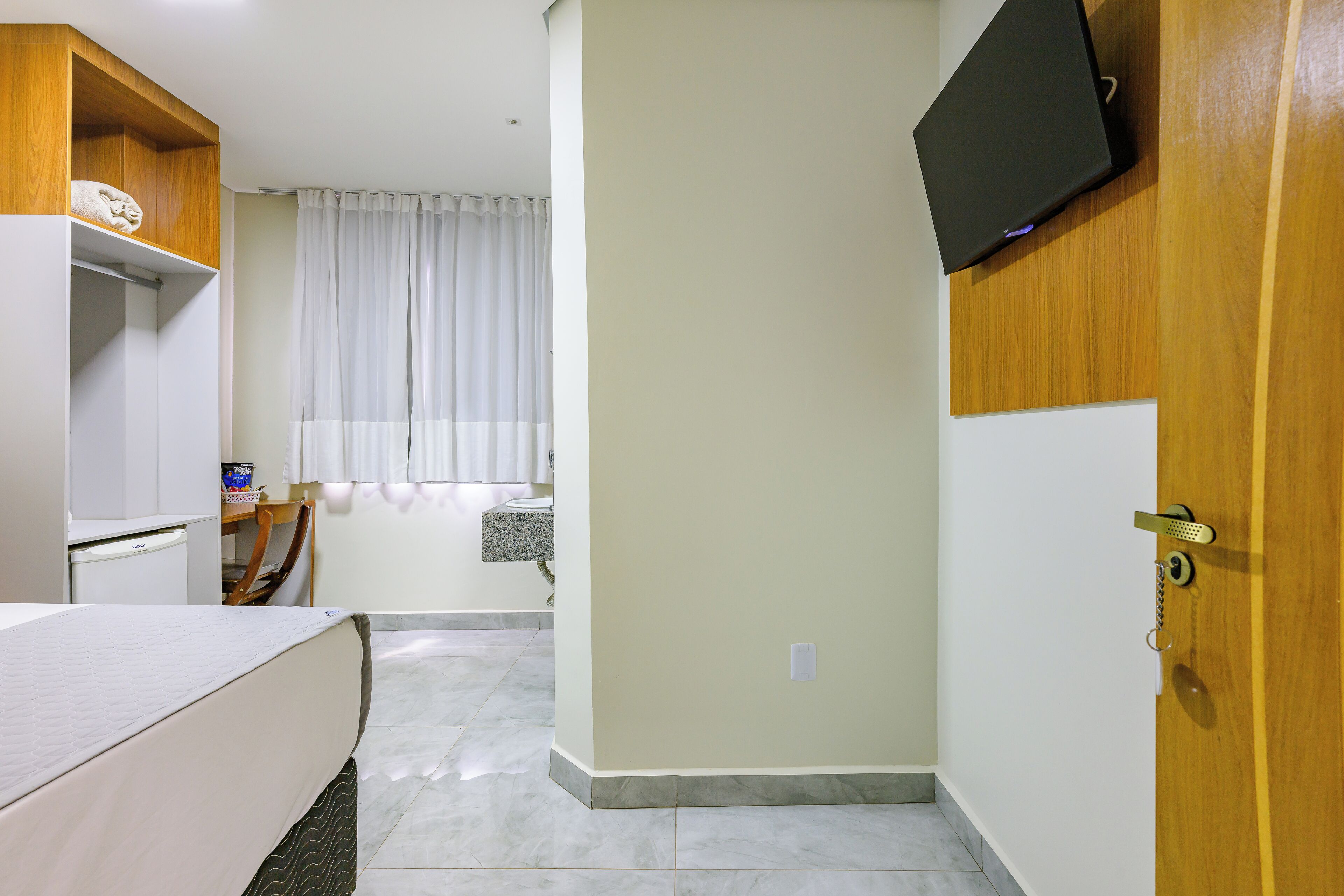 A hotel room with a bed, a desk, a television mounted on the wall, and a window with curtains.