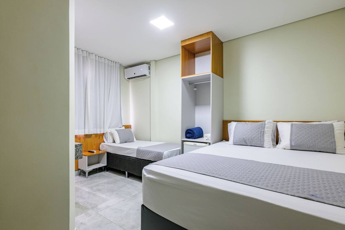 A hotel room with two beds, a wardrobe, a bedside table, an air conditioning unit, and a window with curtains.