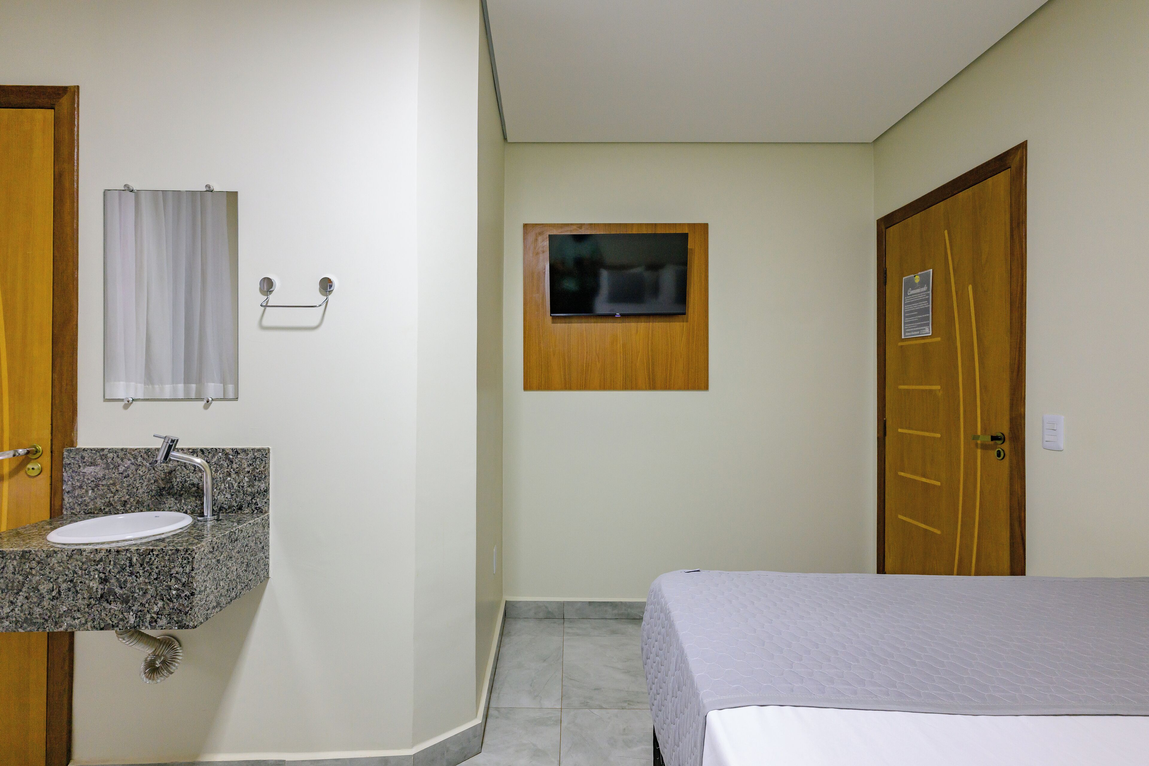A hotel room with a granite countertop sink, a wooden door, a small window, and a bed with a white bedspread.