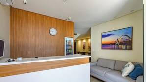 A reception area with a wooden reception desk, a clock, a refrigerator, and a sofa with a painting of a bridge. - OYO Hotel Brisa Tropical, Brasília (São Sebastião)
