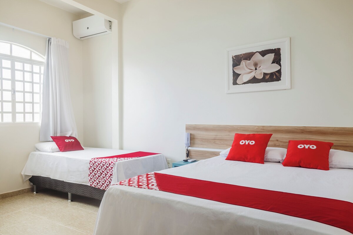 A hotel room with two beds, each with a red and white bedspread and pillows, a wooden headboard, a wall-mounted air conditioner, a window with a white curtain, and a framed picture of a flower on the wall.