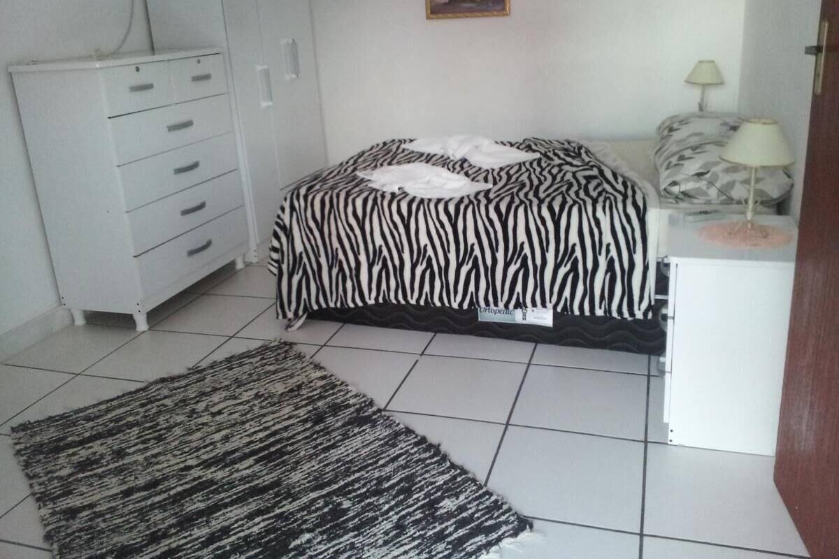 A bedroom with a zebra-patterned bed, a white dresser, a small table with a lamp, and a rug on the floor.