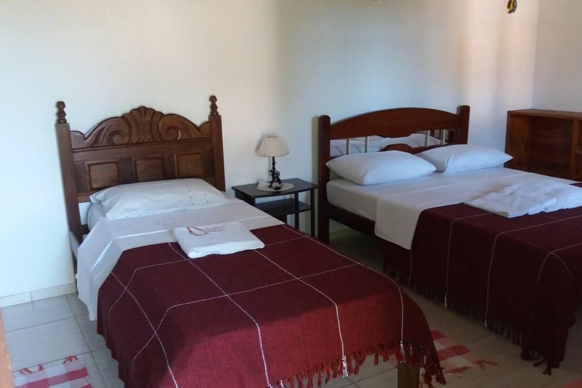 A hotel room with two beds, each with a wooden headboard, white linens, and a red blanket. There is a small bedside table with a lamp and a wooden cabinet.