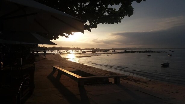 A beach with a wooden bench and a pier in the background. - OYO Pousada Encanto dos Anjos (Búzios)