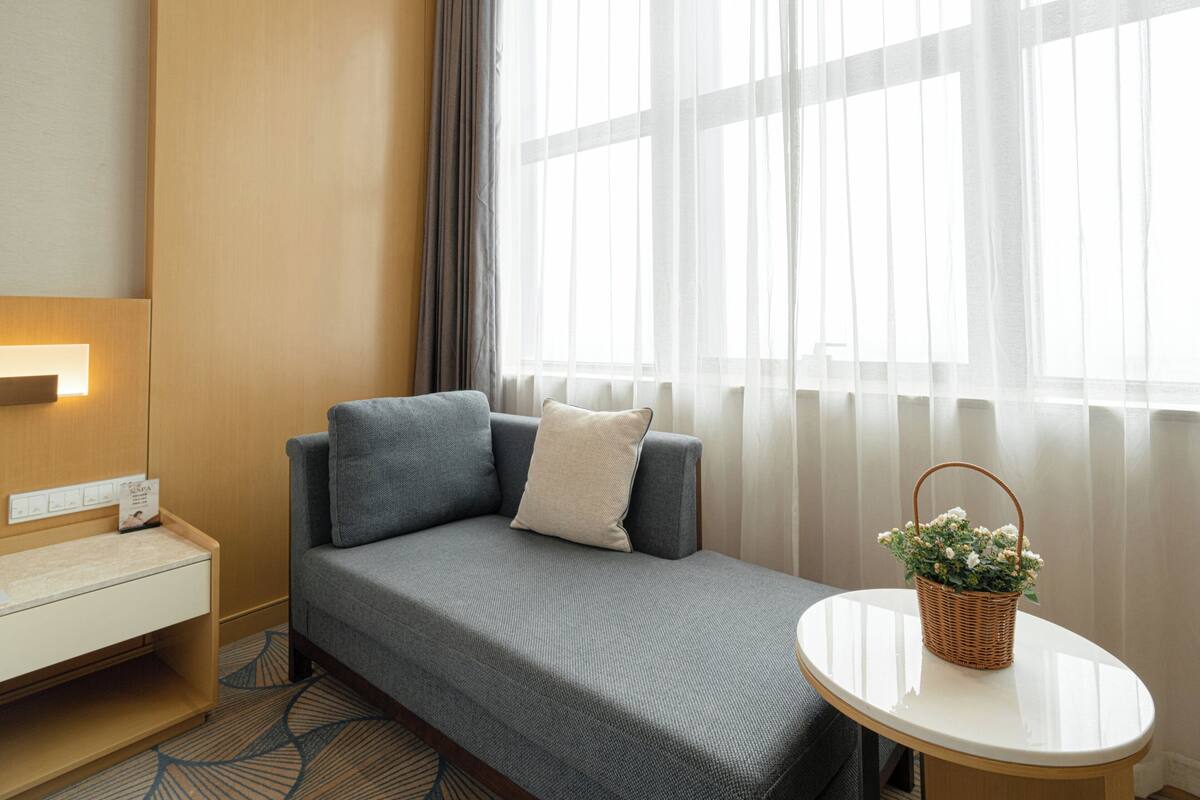 A modern hotel room with a grey sofa, a small bedside table, a white round table, and a basket of flowers.