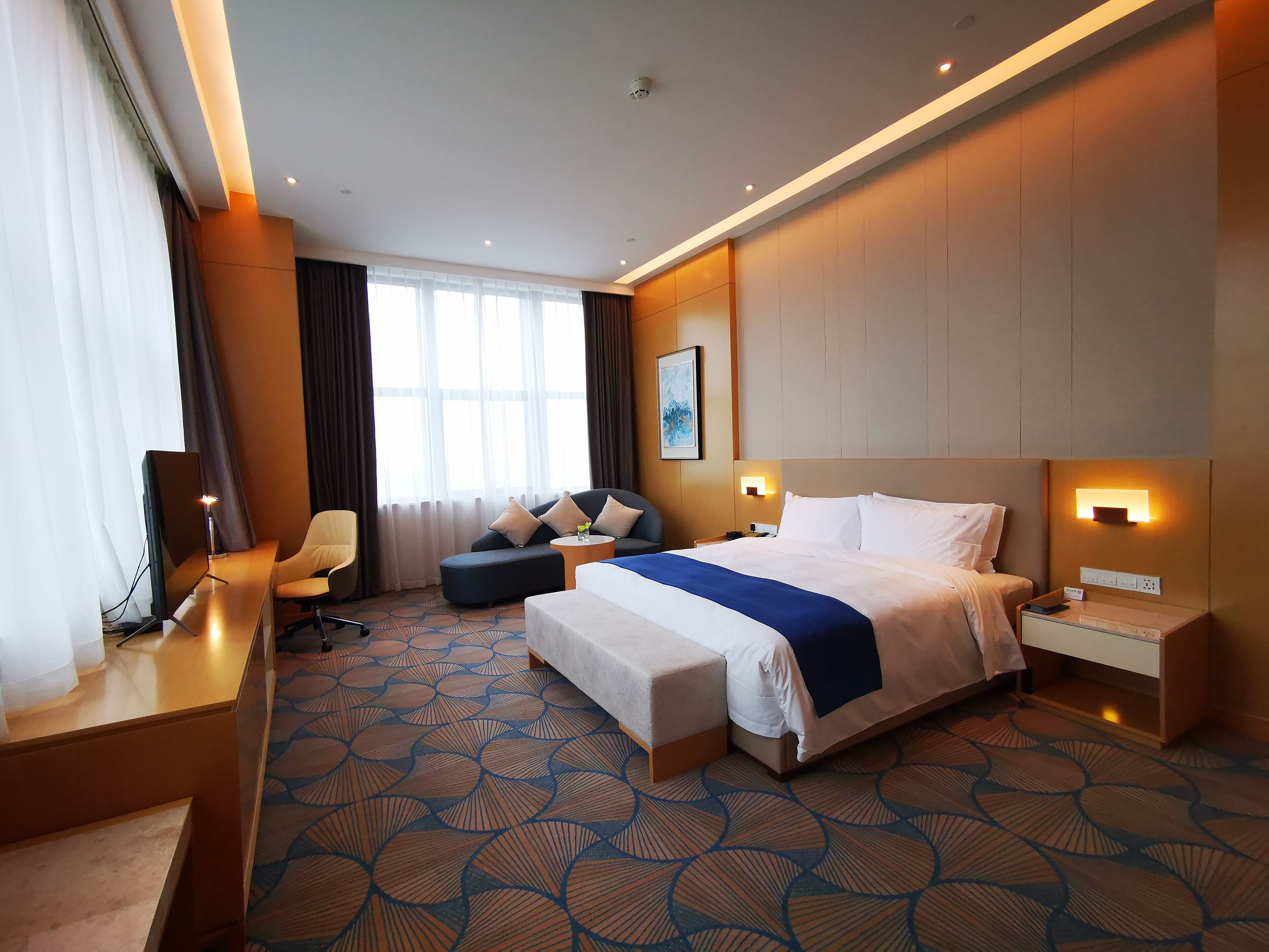 A hotel room with a large bed, a sofa, a desk with a computer, and a nightstand with a lamp.