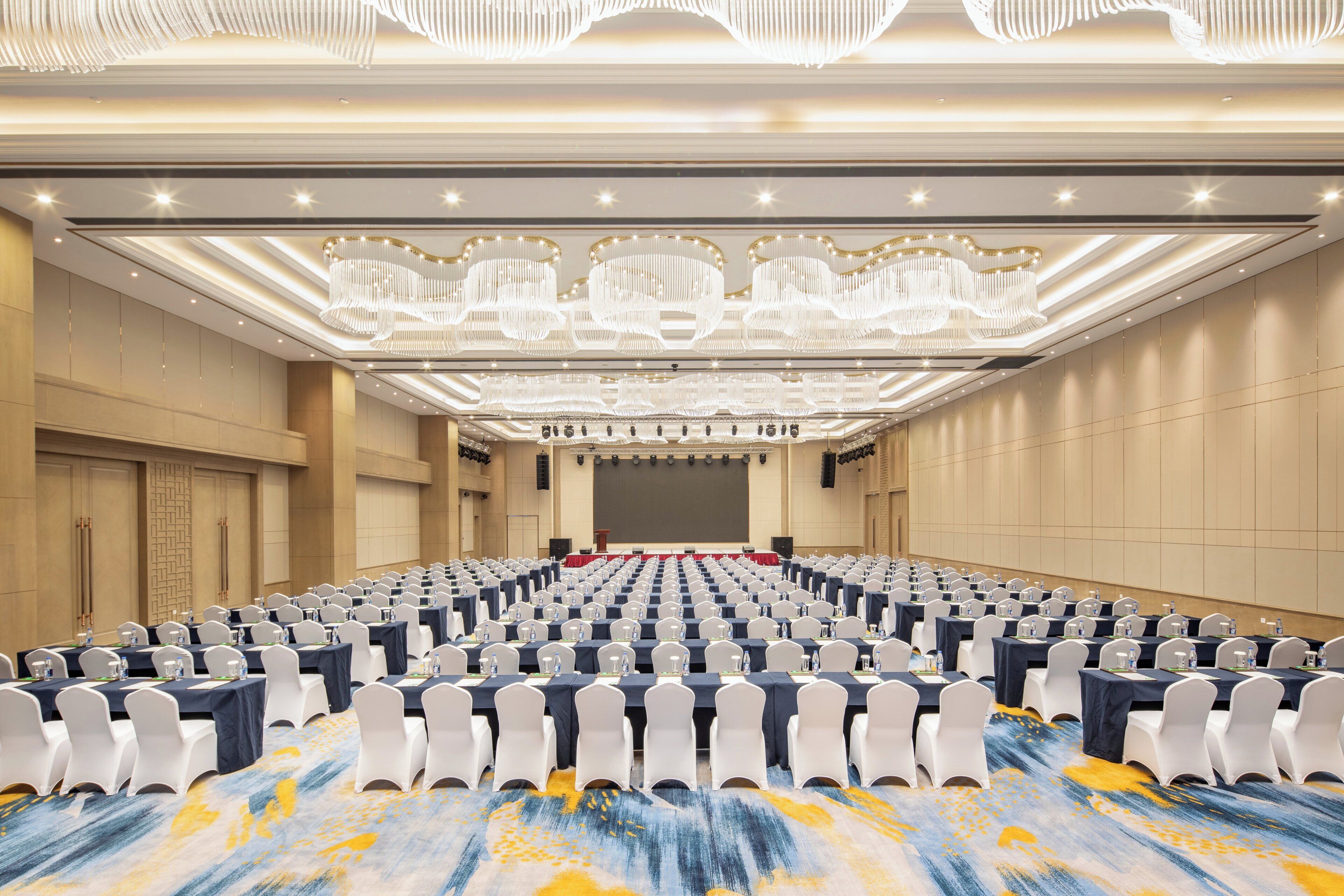 A large conference room with rows of tables and chairs, a stage with a red carpet, and a ceiling with ornate chandeliers.