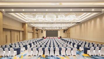 A large conference room with rows of tables and chairs, a stage with a red carpet, and a ceiling with ornate chandeliers.