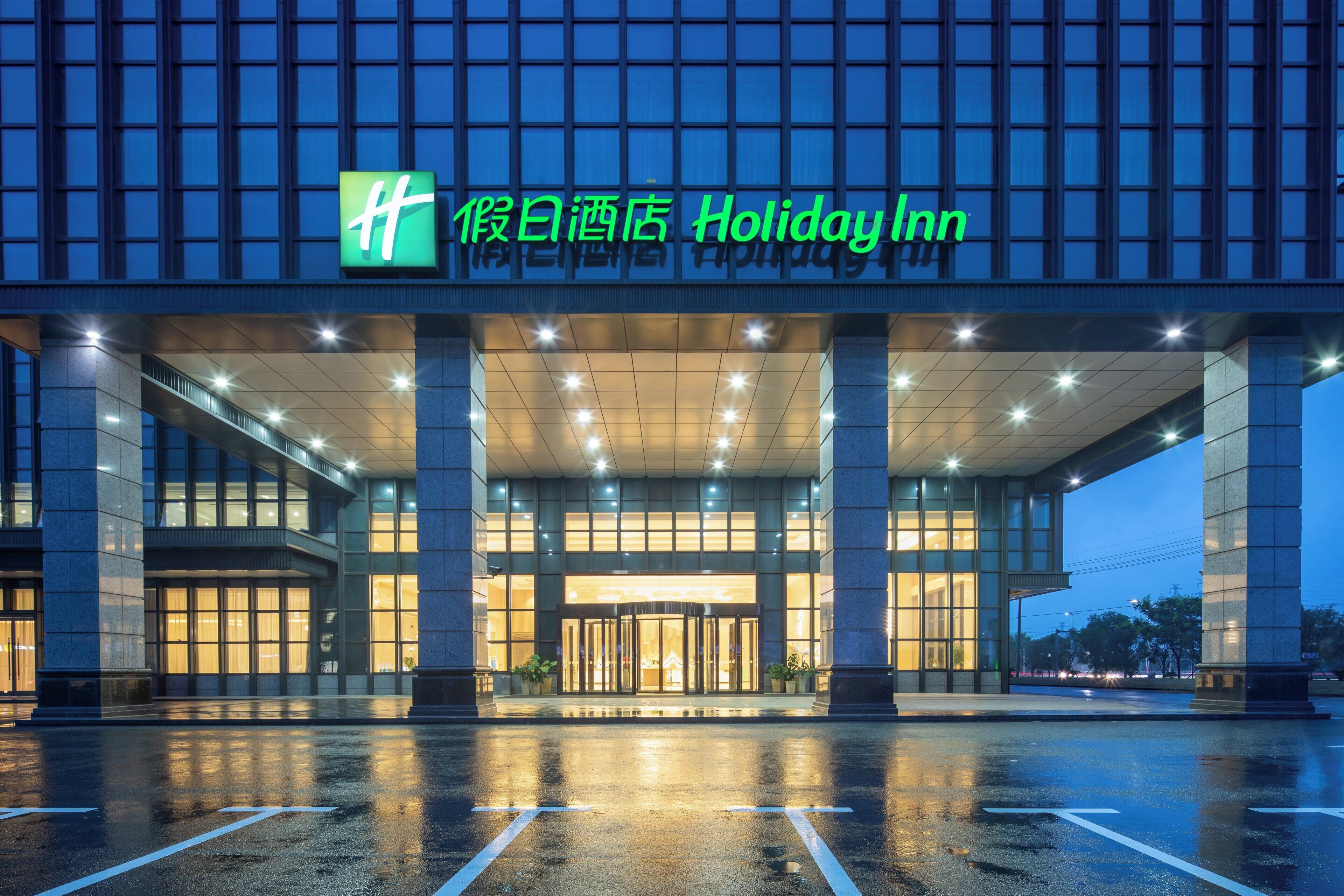 Entrada de un hotel Holiday Inn con fachada de vidrio, señalización iluminada y pavimento mojado.