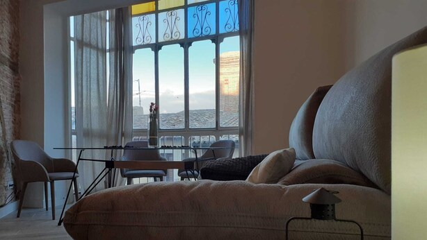 Un salon chaleureux avec un grand canapé, une petite table sur laquelle est posée une bougie, et une vue sur le paysage urbain à travers une fenêtre ornée de vitraux.