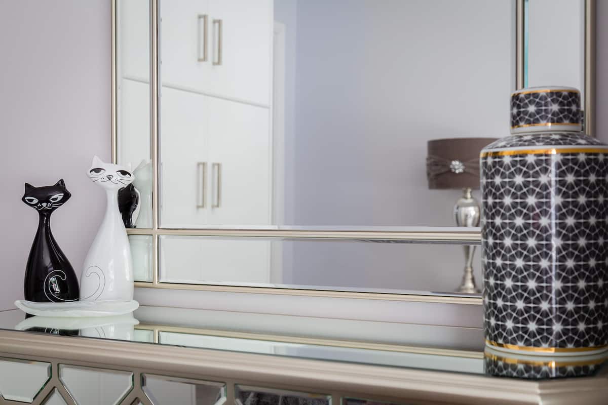 Une coiffeuse avec un miroir, une statuette de chat noir et blanc, un vase blanc et un récipient à motifs.