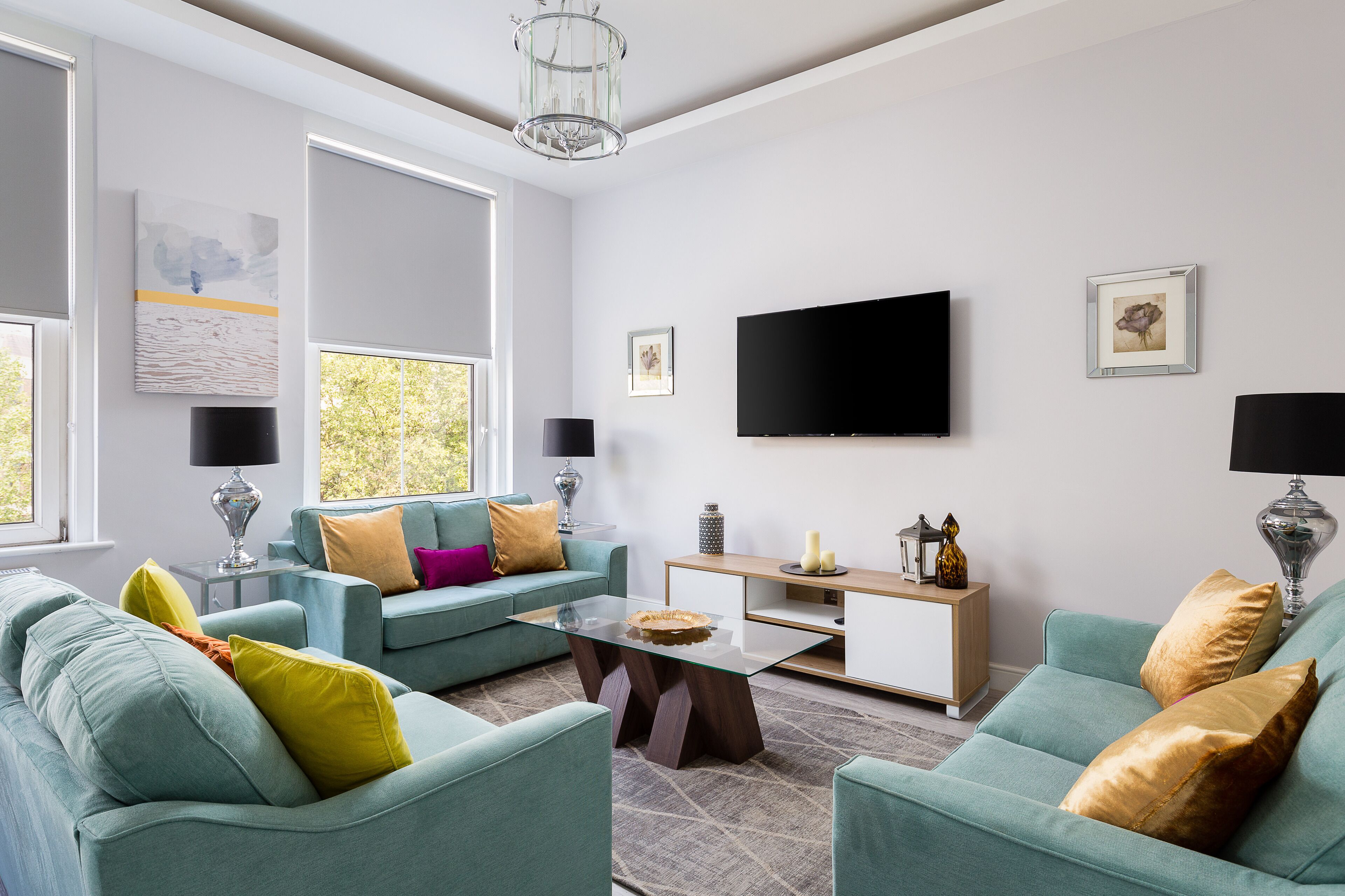 A modern living room with teal sofas, a glass coffee table, and a flat-screen TV mounted on the wall.
