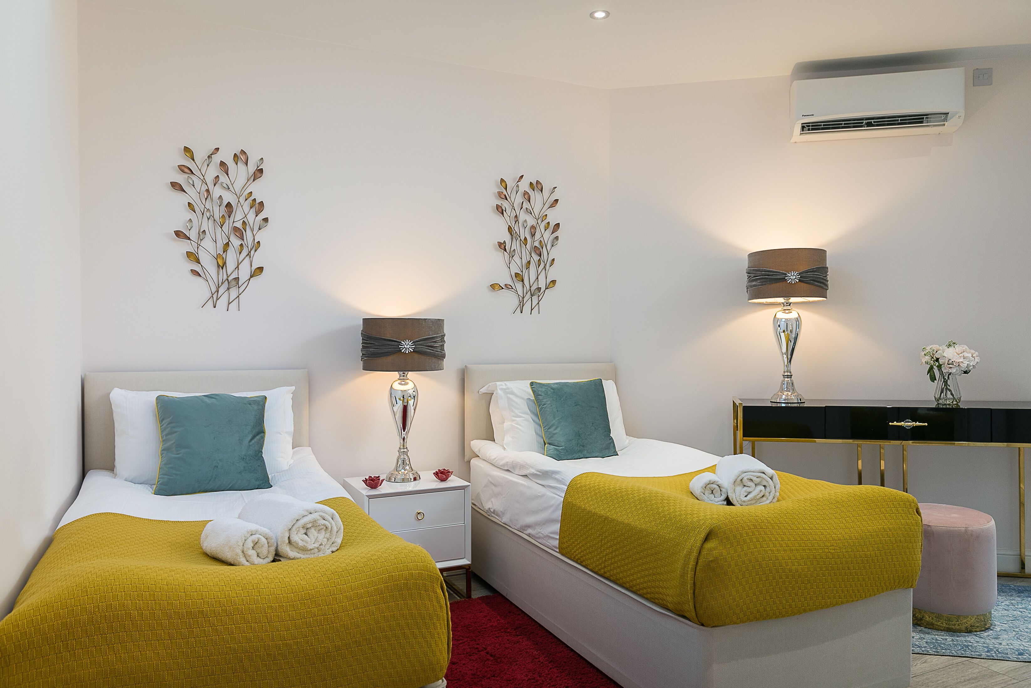 Habitación de hotel con dos camas, cada una con un edredón amarillo y almohadas blancas, una mesita de noche con una lámpara y decoración en la pared.