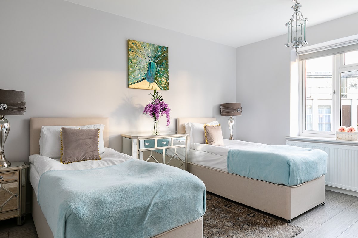 Une chambre d’hôtel avec deux lits, une table de chevet avec une lampe, une commode, un tableau représentant un paon et une fenêtre donnant sur un beau paysage.