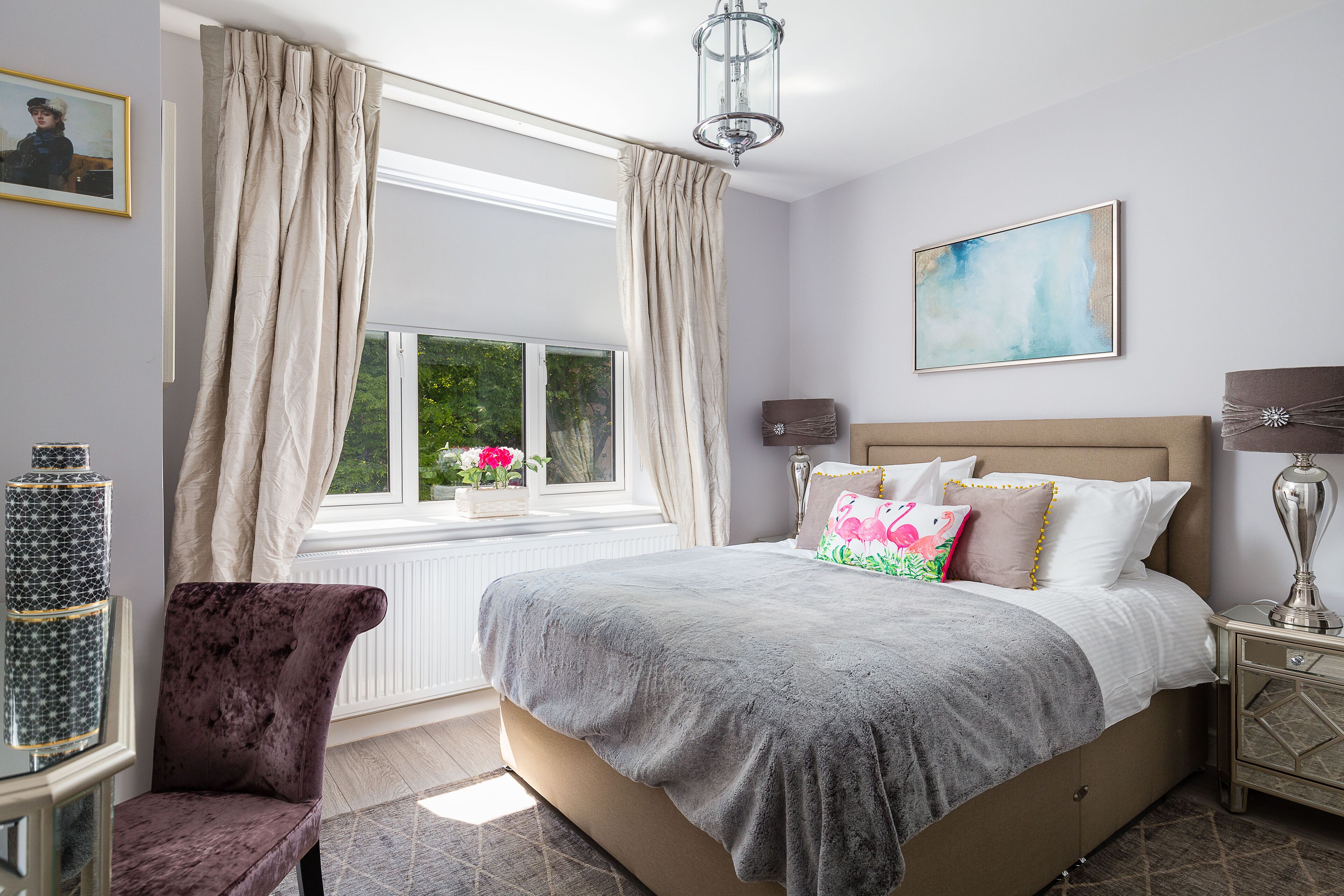 A bedroom with a large bed, a window with curtains, a bedside table with a lamp, and a chair.