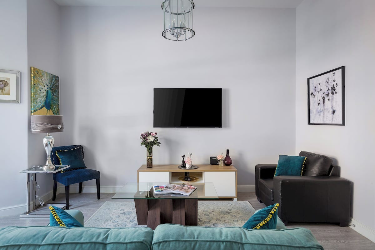 Un salon moderne avec une table basse en verre, un fauteuil bleu, un canapé noir, une console blanche et un téléviseur à écran plat fixé au mur.