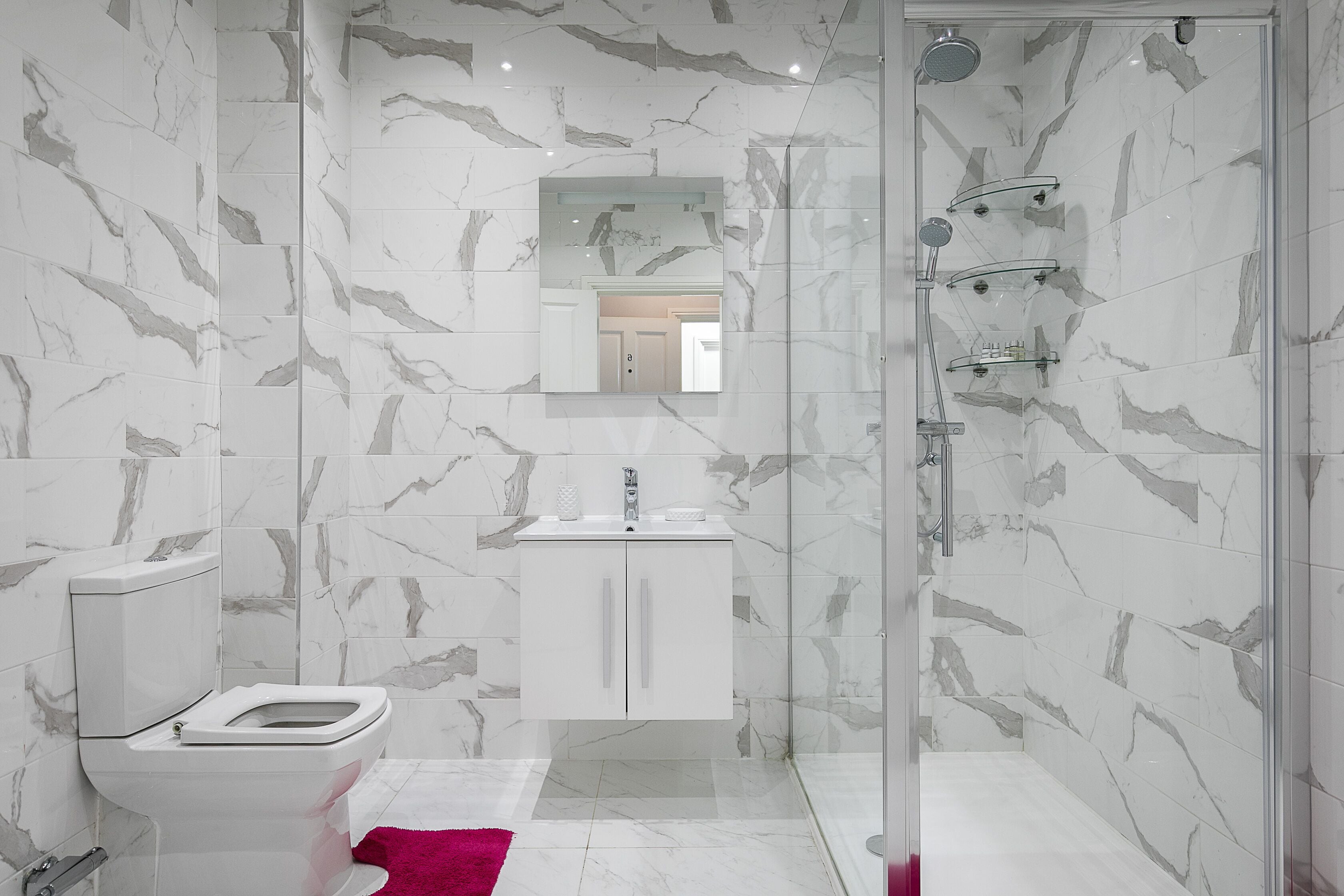 Un baño moderno con paredes de mármol, ducha envuelta en vidrio, inodoro blanco y un tocador pequeño.