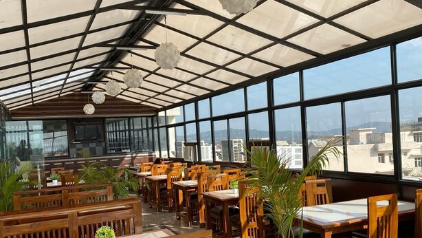 A restaurant with wooden tables and chairs, large windows, and a view of buildings. - Hotel Royal Stay (Reasi)