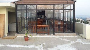 A covered outdoor seating area with glass walls, wooden furniture, and a potted plant. - Hotel Royal Stay (Reasi)