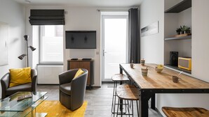 Una sala de estar moderna con una mesa de centro de vidrio, sillas de cuero, una mesa de comedor de madera con taburetes altos y un televisor de pantalla plana, además de un amplio ventanal con cortinas.