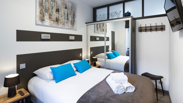 Una habitación de hotel moderna con una cama grande, una mesita de noche con lámpara, un espejo y un televisor montado en la pared.