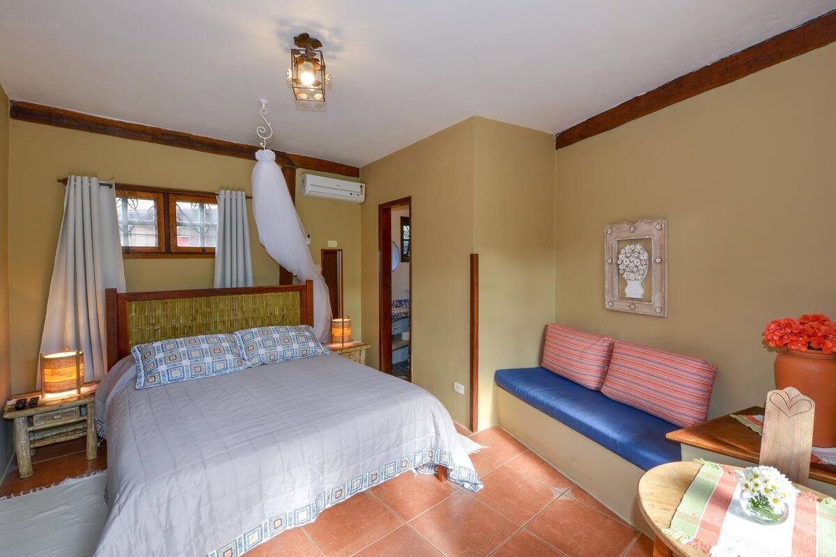 Une chambre à coucher comprenant un lit, des lampes de chevet, une table de chevet, un banc avec des coussins, un luminaire fixé au mur et un tableau encadré.