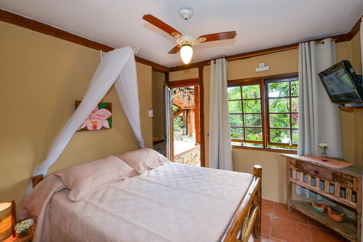 Une chambre à coucher comprenant un lit, un ventilateur de plafond, une télévision, une fenêtre avec des rideaux et une armoire en bois.