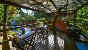 An outdoor wooden deck with tables and chairs, surrounded by lush greenery and a pool. - Pousada Villa da Prainha (Ilhabela)