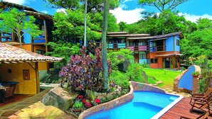 A pool area with a wooden deck, surrounded by colorful buildings and lush greenery. - Pousada Villa da Prainha (Ilhabela)