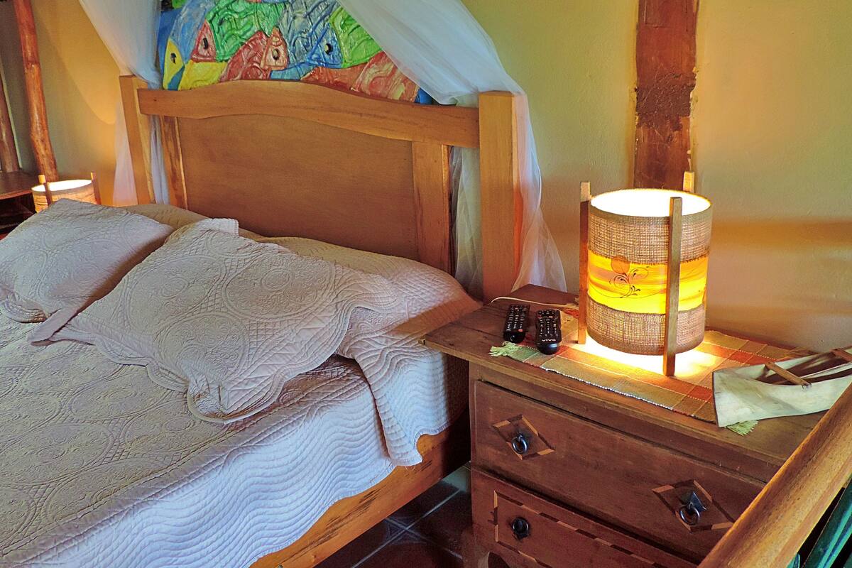 Un lit en bois avec un baldaquin, une lampe de chevet, deux télécommandes et une petite table de chevet en bois dotée de tiroirs.