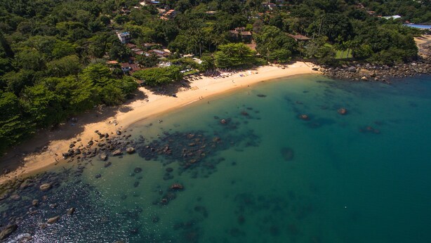 An aerial view of a beach with clear turquoise waters, surrounded by dense greenery and scattered houses. - Pousada Villa da Prainha (Ilhabela)