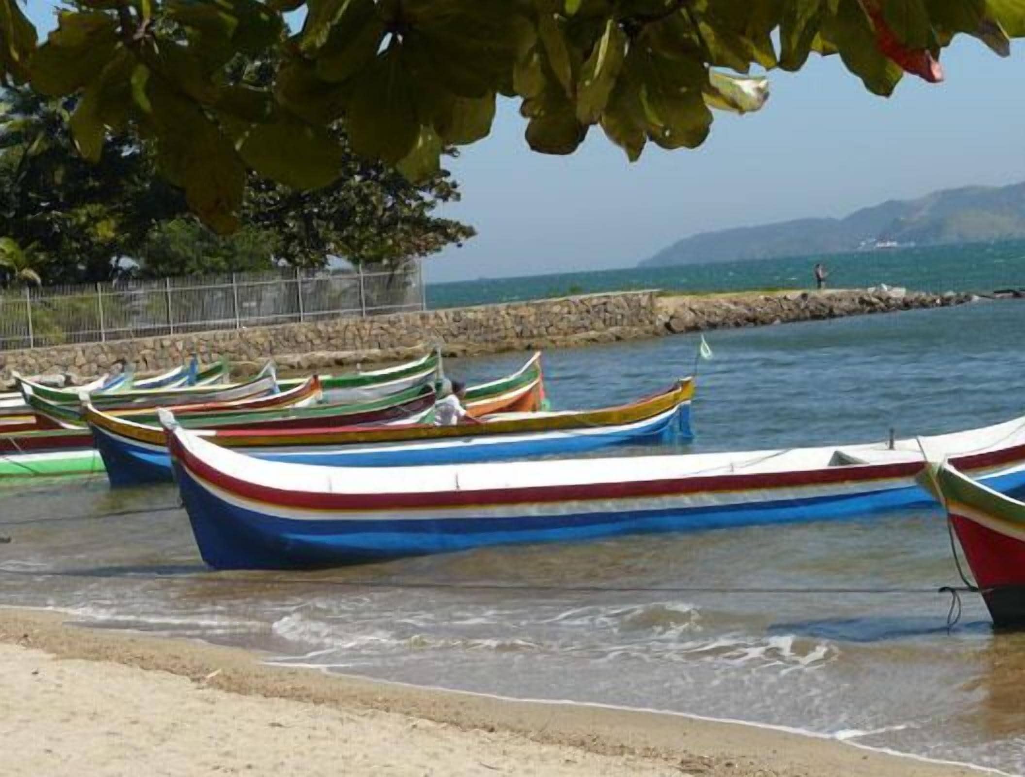 Uma fileira de barcos coloridos atracados em uma praia, com um muro de pedra e uma vista desobstruída do mar e montanhas ao longe.