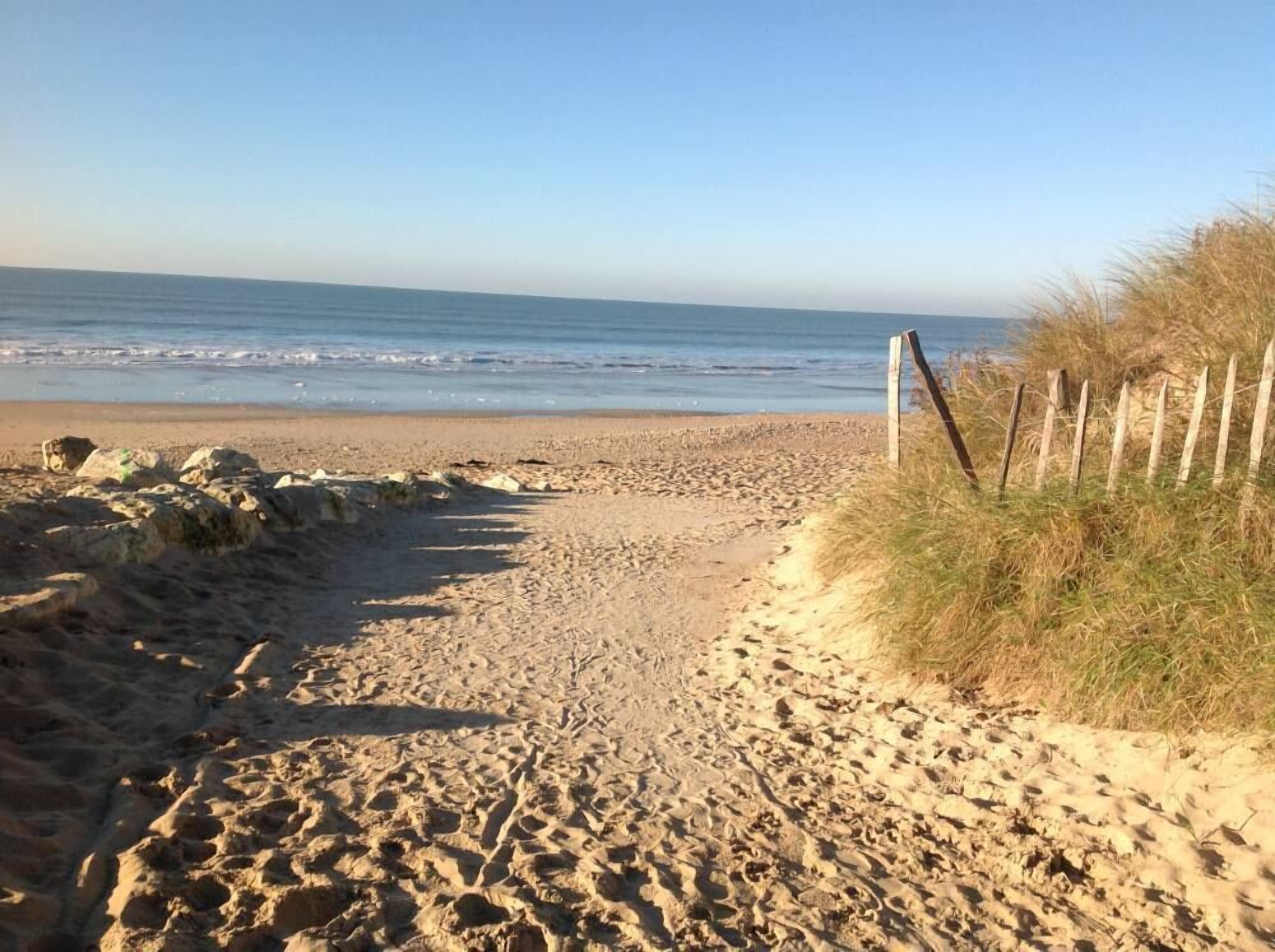 Een zandpad langs het strand dat naar de oceaan leidt, met een houten hek aan de rechterkant en rotsen aan de linkerkant.