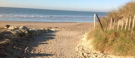 Een zandpad langs het strand dat naar de oceaan leidt, met een houten hek aan de rechterkant en rotsen aan de linkerkant.