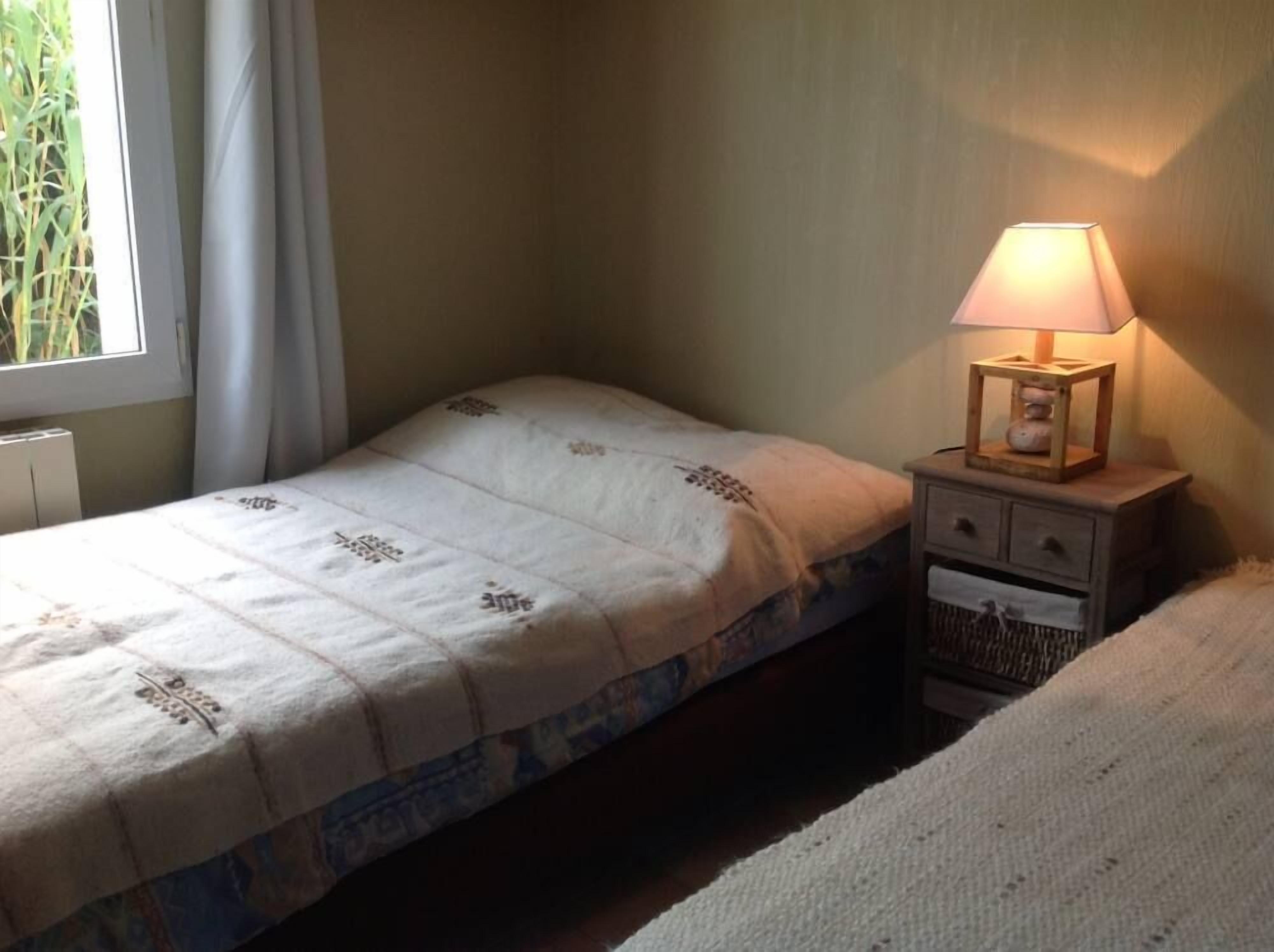 Ein kleines, schlichtes Zimmer mit einem Einzelbett, einem Nachttisch mit Lampe und einem fenster mit Vorhängen.