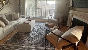 A living room with a sofa, ottoman, chairs, a patterned rug, a fireplace, and a large window with a view of trees. - Sugarbush Mountainside Retreat - Ski in ski out (Warren)