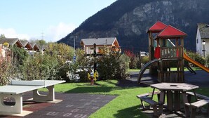 A playground with a slide and climbing structure, surrounded by houses and a mountain in the background. - Petit Nid Douillet au Coeur de Salvan (Salvan)