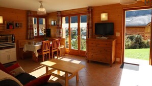 A cozy living room with a wooden table, chairs, and a sofa. There's a television on a wooden cabinet, and a kitchen area with a table and chairs. - Petit Nid Douillet au Coeur de Salvan (Salvan)