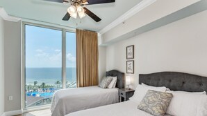 Een hotelkamer met twee bedden, een plafondventilator en een groot raam met uitzicht op de oceaan en een resort.