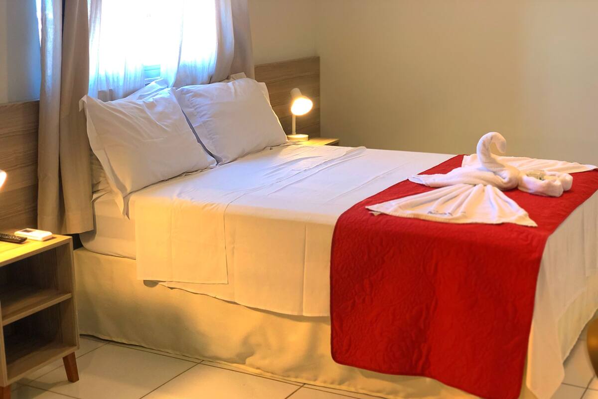 A neatly made bed with white linens and a red blanket, a bedside lamp, and a small wooden nightstand with a remote control.