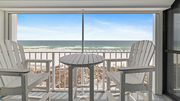 A balcony with two white Adirondack chairs and a small round table, overlooking a beach with the ocean and a clear sky.