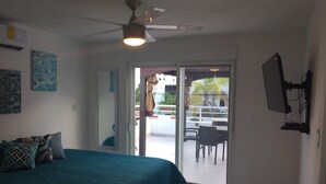 Une chambre avec un lit, un ventilateur de plafond et une porte-fenêtre coulissante donnant sur une terrasse extérieure équipée d’une table et de chaises.