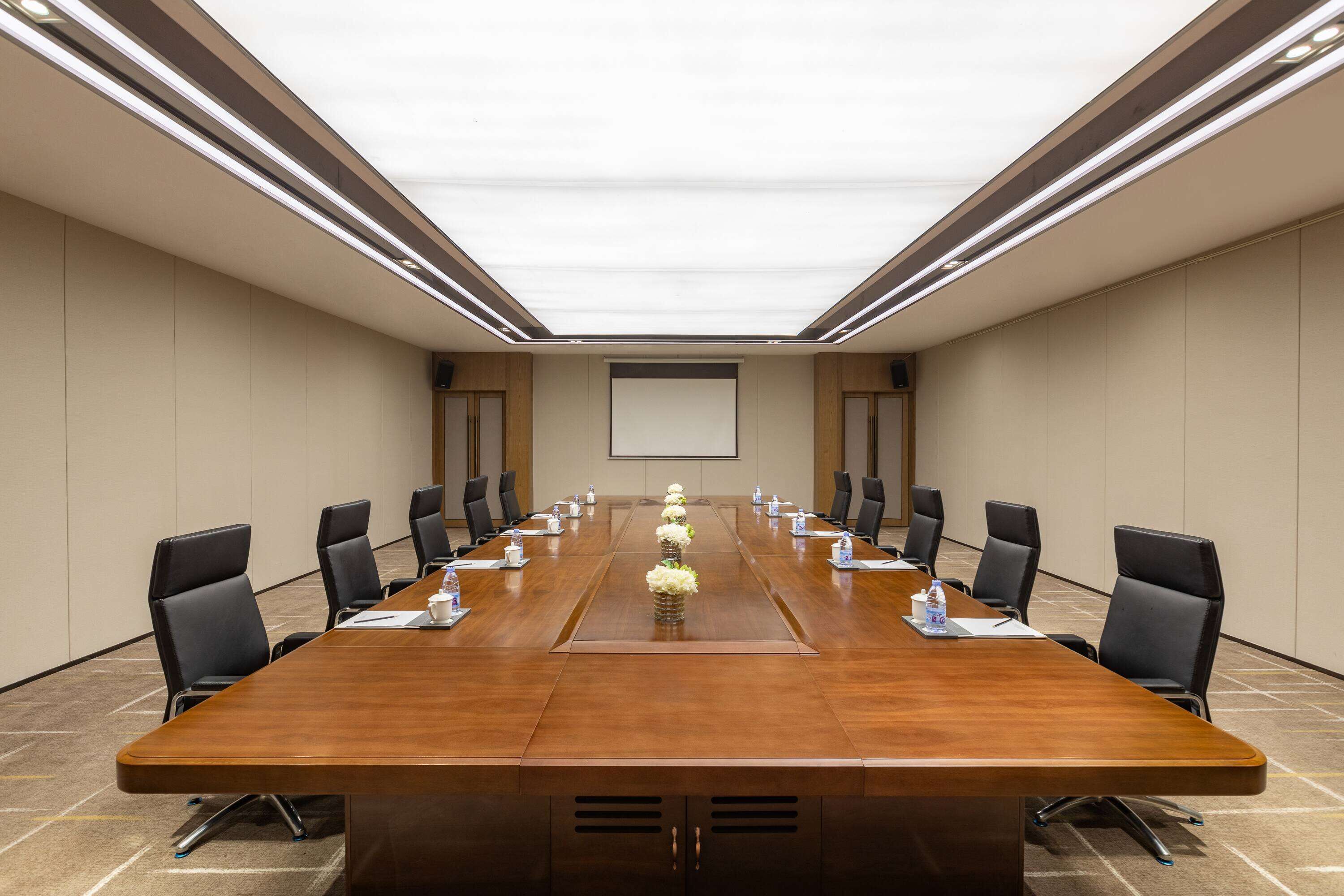 Un salón de conferencias con una larga mesa de madera, sillas negras y una pantalla de proyector al frente.