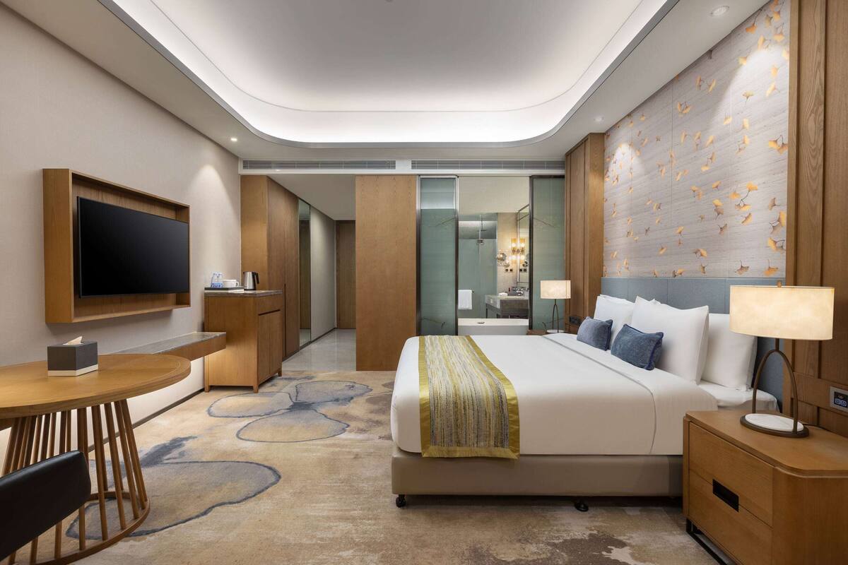 Una habitación de hotel moderna con una cama grande, mesitas de noche, televisión y un baño visible a través de una puerta de cristal.