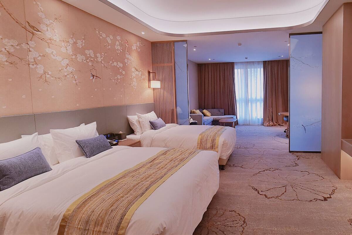 Habitación de hotel con dos camas, zona de estar, televisión y un amplio ventanal con cortinas.