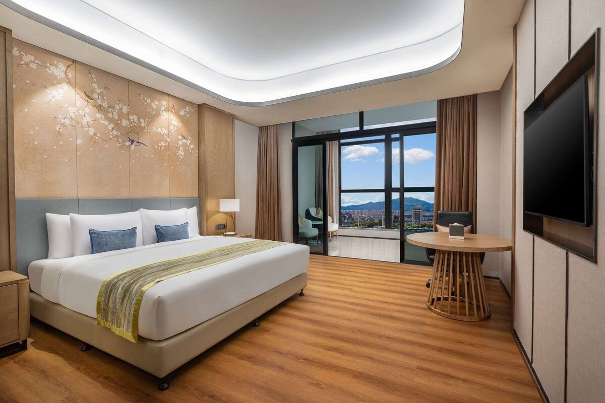 Habitación de hotel moderna con una cama grande, un escritorio, un televisor y un balcón con vista a la ciudad.