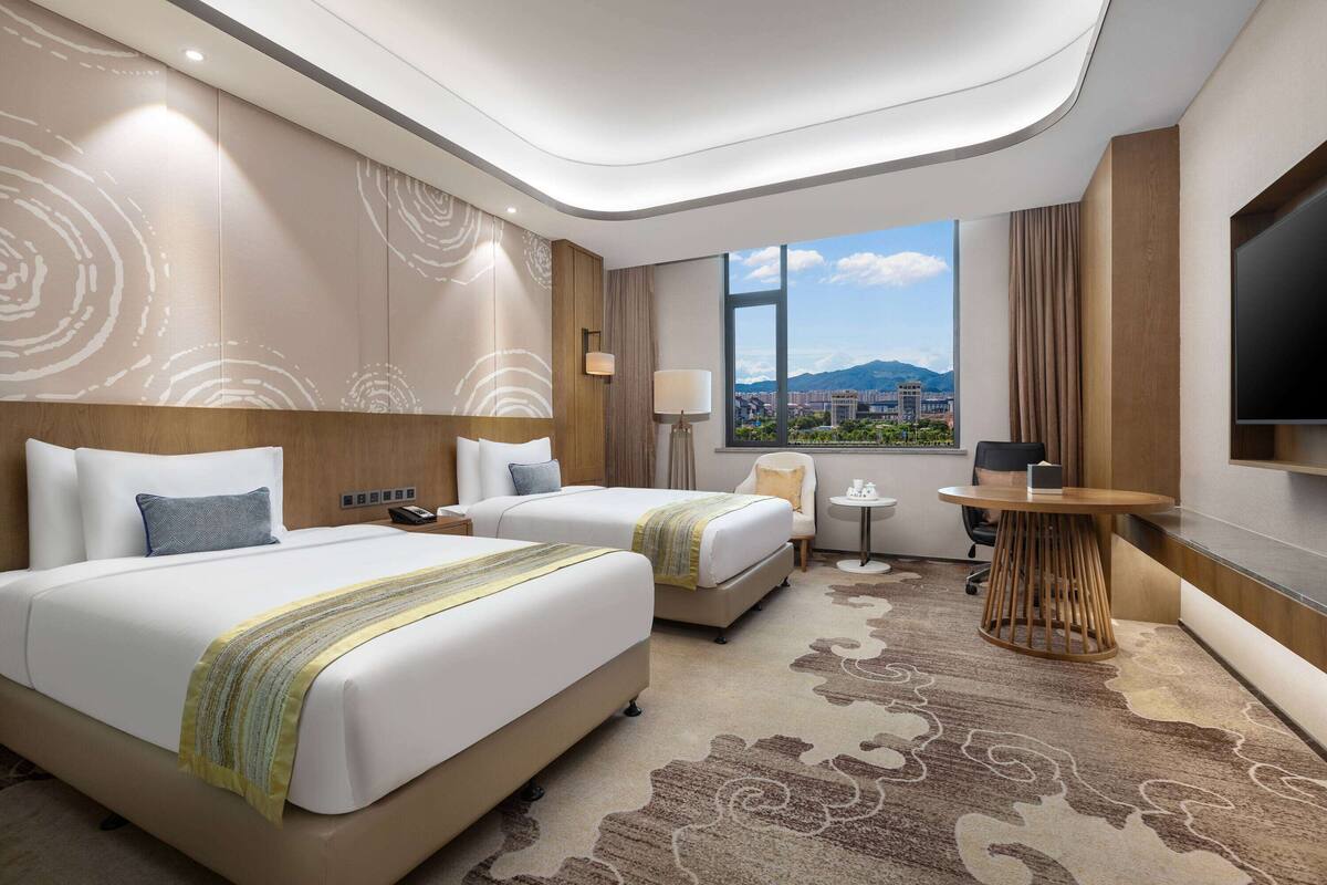 Habitación de hotel con dos camas, un escritorio, una silla, un televisor y un ventanal con vista a la ciudad.