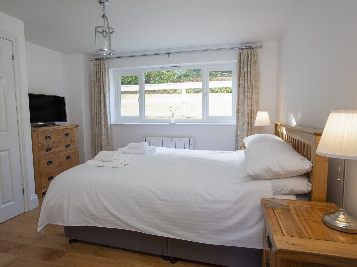Un dormitorio con cama, cómoda, televisor, ventana con cortinas y lámpara.