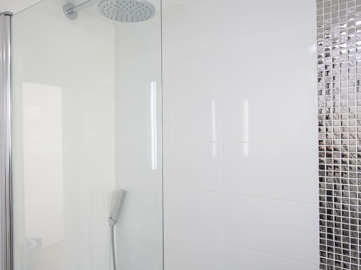 Una ducha moderna con puerta de vidrio, cabezal de ducha de mano y pared de azulejos con un mosaico decorativo.